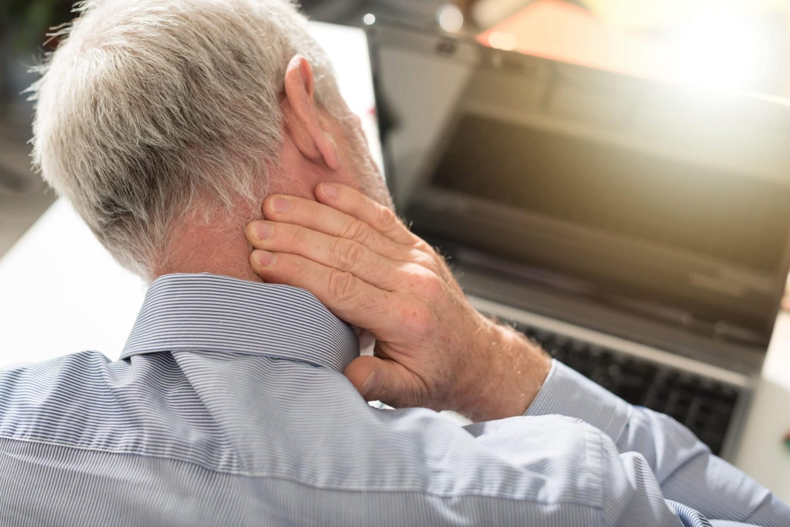 Head-Neck-Masses-evaluation Older man looking down at a computer has his hand on that back of his neck that is in pain.